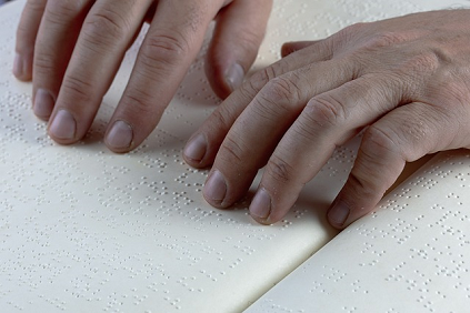 A close-up of a pair of hands resting on an open book with fingertips reading a page of braille.