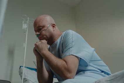 a middle-aged man in a hospital gown sits on a hospital bed, leaning forward with his hands clasped together and his chin resting on them. He stares down at the ground, appearing deep in thought.