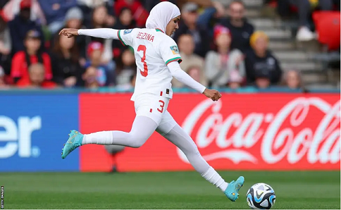 "Moroccan defender Nouhaila Benzina looks down at the ball during a Women's World Cup match, right leg drawn back to kick the ball in front of her. She wears an all-white kit with her surname and the number 3 on the back, white leggings, a white hijab and light blue boots. A crowd watches in the background."